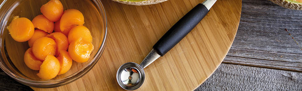 Two cantaloupe melon halves with melon baller tool and bowl of melon balls on round cutting board