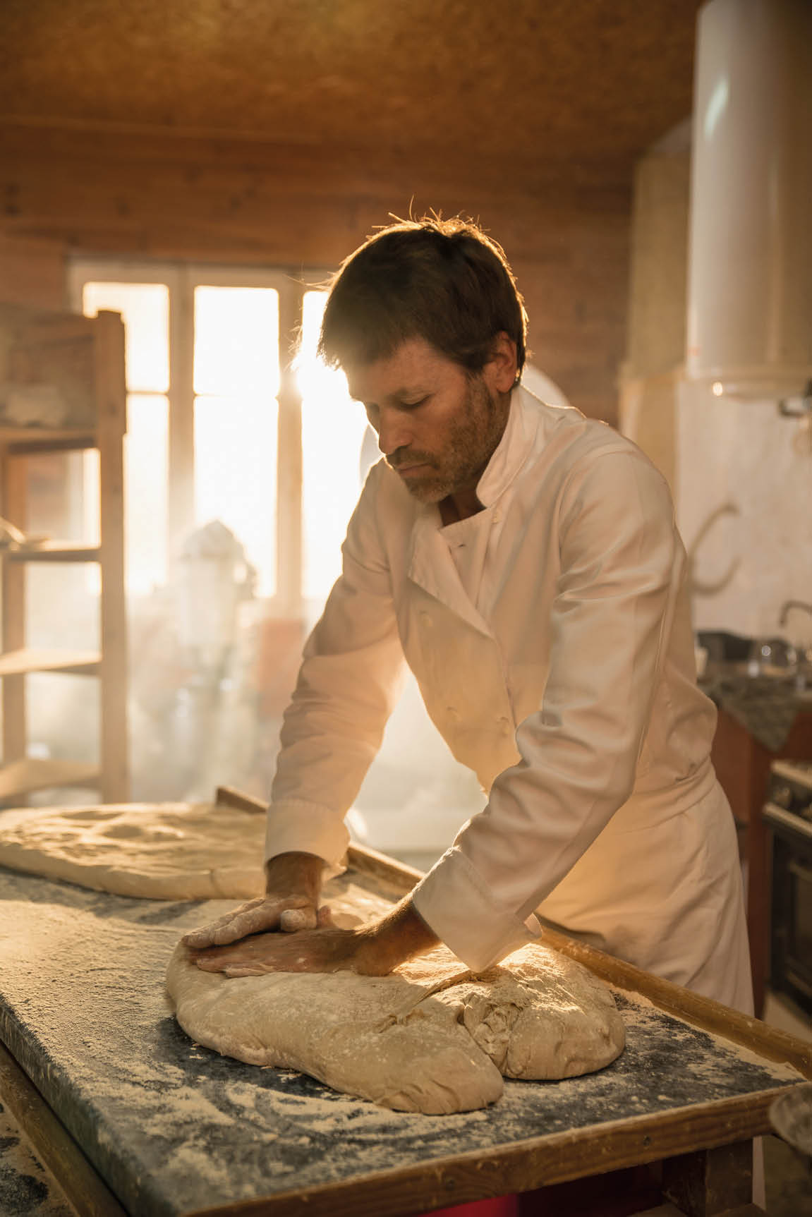 in an artisan bakery, the baker kneads the dough for the bread. The morning sun comes in through the window