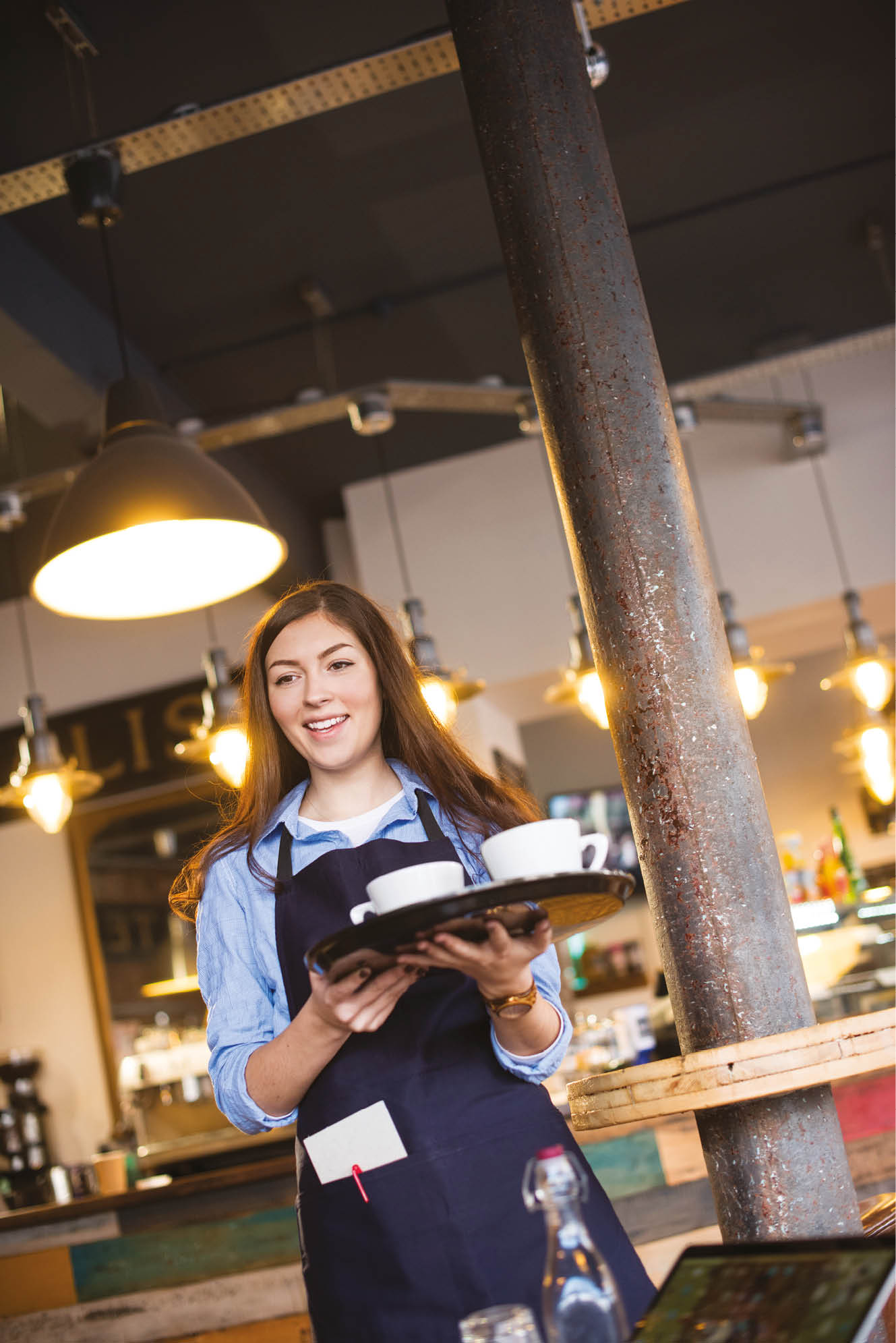 young coffee shop waitress walks towards a customer a trendy warehouse style cafe . She is wearing a blue apron and carrying a tray with the customers drink on.