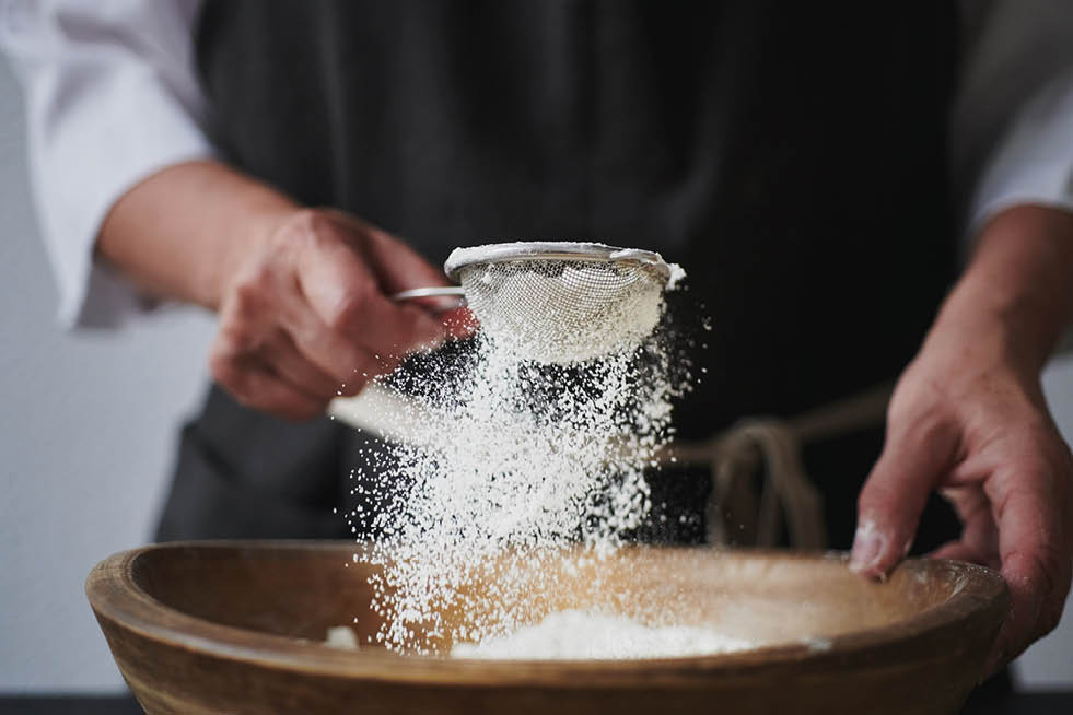 Slow motion shot of aged female hands sifting flour by sieve in wooden bowl.